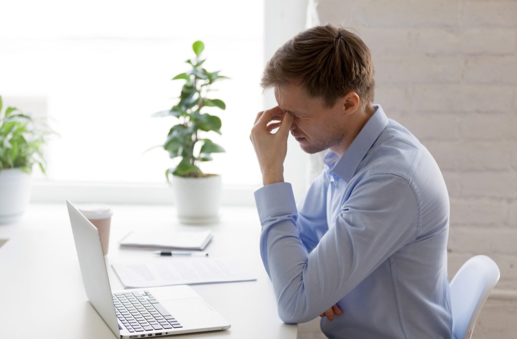 A young adult man rubbing his eyes due to discomfort while using a computer, which is an activity that can exacerbate symptoms of Dry Eye Syndrome.