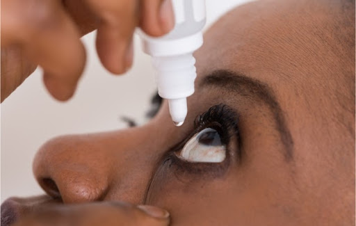 A drop of solution hangs from a small bottle of eye drops as as young woman holds it while she applies them to her irritated eye.