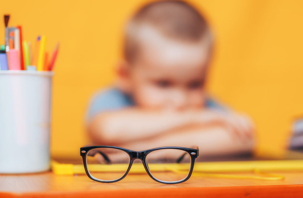 A boy sitting behind a pair of glasses placed on the table.