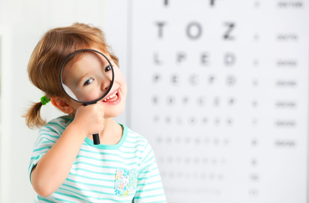 Young smiling girl holding a magnifying glass up to her face and sitting in front of a blurred out eye chart in the background.