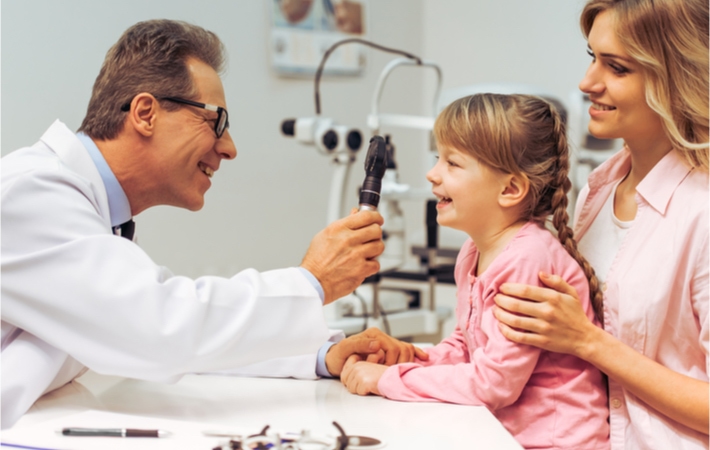 Smiling mature optometrist examines a smiling young girl with an ophthalmoscope as her hold helps to hold her from behind.
