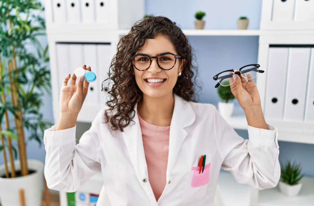 Smiling female optometrist holding up a pair of glasses in her left hand and a contact lens case in her right hand.