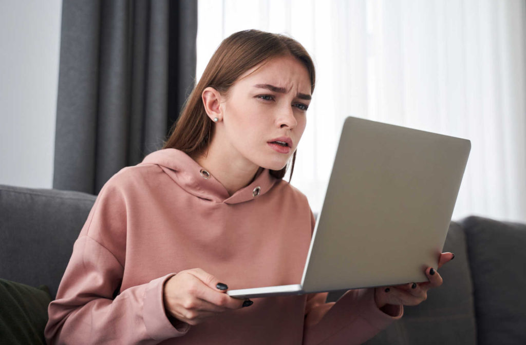 A young girl student sitting on a sofa is squinting while looking on the screen of her laptop.