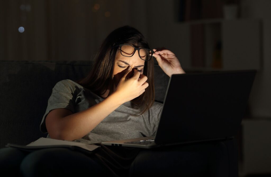 A woman experiencing eye strain while working using her laptop in a dark room.