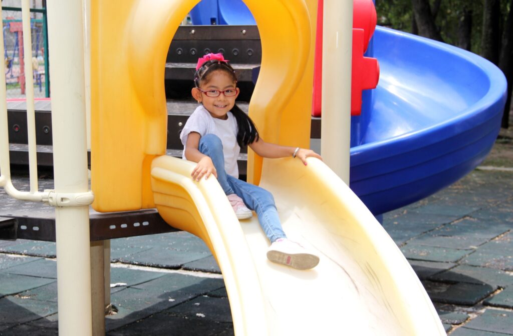 Young girl wearing glasses and a pink headband smiling while sliding down a yellow playground slide on a sunny day.