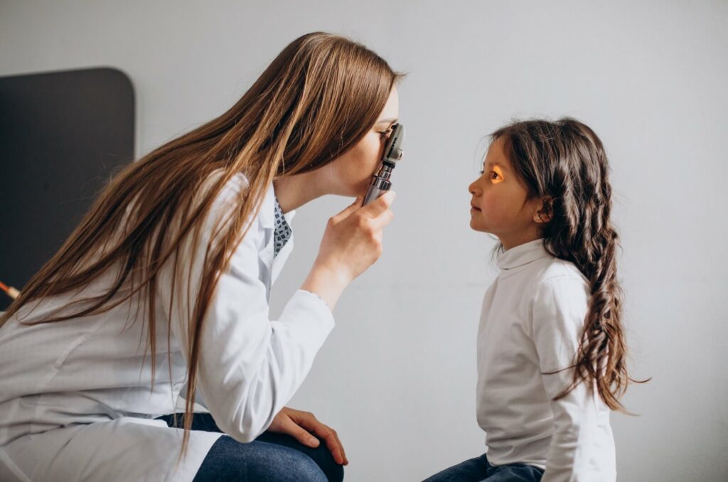 A female optometrist in a white coat examines a young girl's eyes using an ophthalmoscope. The girl, dressed in a white turtleneck, sits still and looks straight ahead while the eye examination takes place in a well-lit room