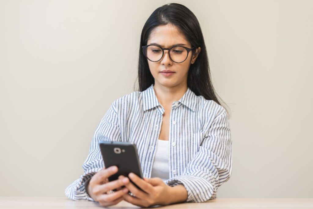 Young woman in striped shirt wearing eyeglasses while looking at smartphone against neutral background.