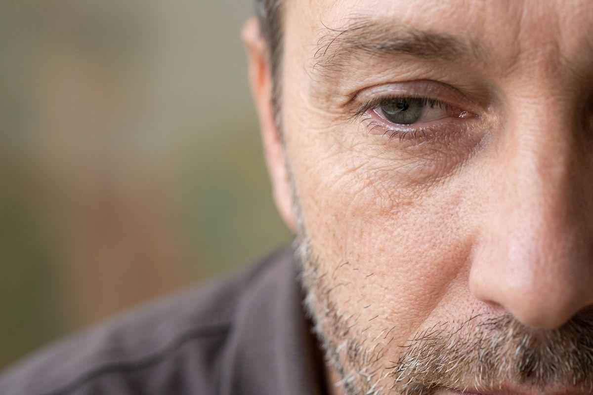 Close-up of half an adult's face with a grey stubble beard, looking down with a serious expression against a blurred background.